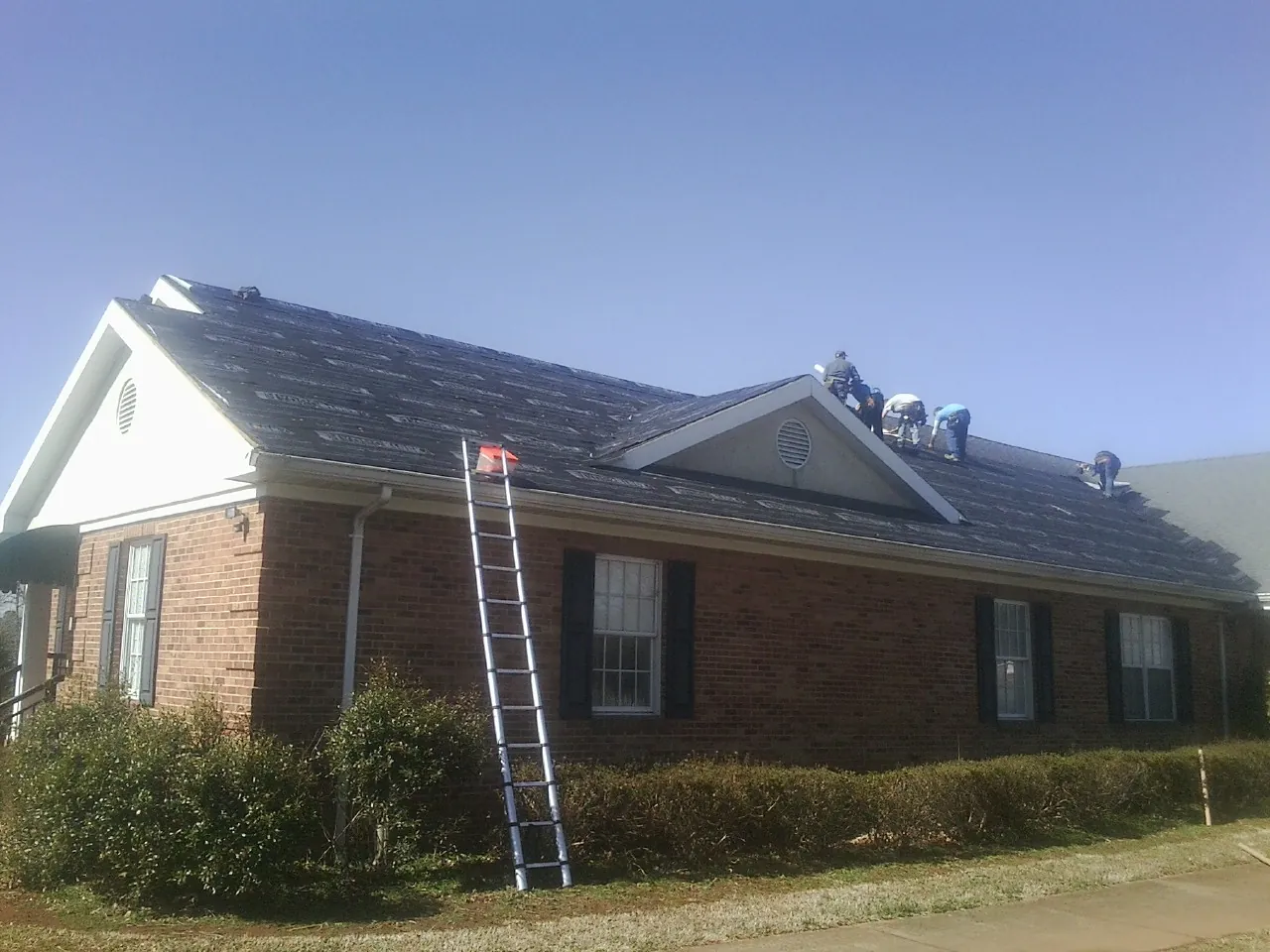 Crew tearing shingles off a brick ranch roof with a ladder set — Emergency Roof Repair work by our crew in Fingerville