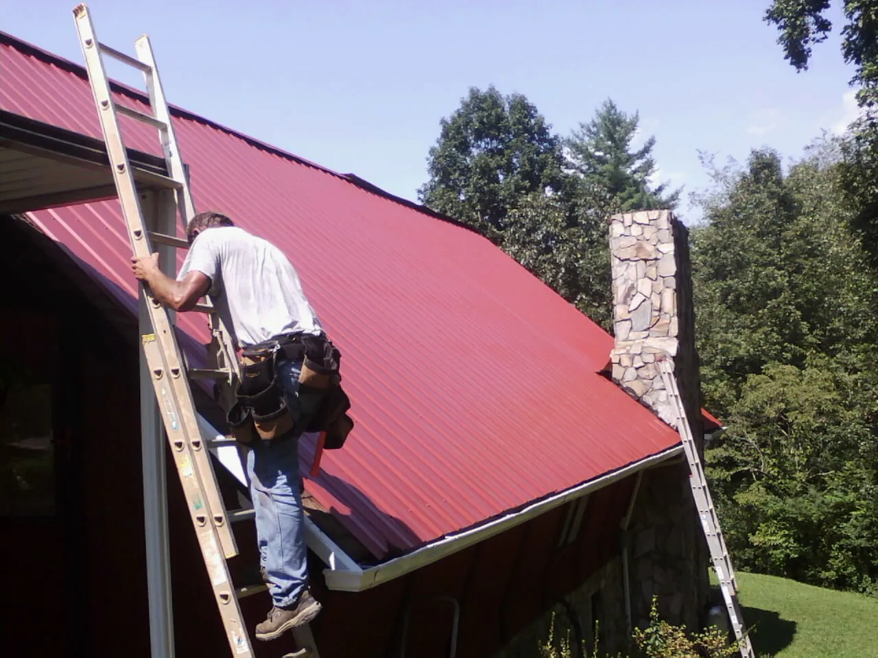 Roofer on a ladder beside a red metal roof and stone chimney — Soffit & Fascia Repair by Roofing Brothers in Fingerville